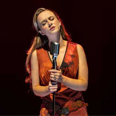 A small white woman with curly hair wearing a red dress sings into an old-fashioned microphone. Red lights bulbs are visible in the background.