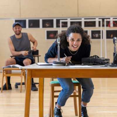 Anoushka Lucas, one of the shows leads is sitting at a table. She is wearing a black jumper and blue jeans. She is leaning forward over the table laughing