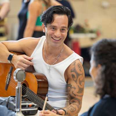 Lead actors Ramin Karimloo and Anoushka Lucas are sat at a table, speaking with eachother. Ramin is smiling and we see Anousha's face at a 3/4 angle. She appears to be smiling back.