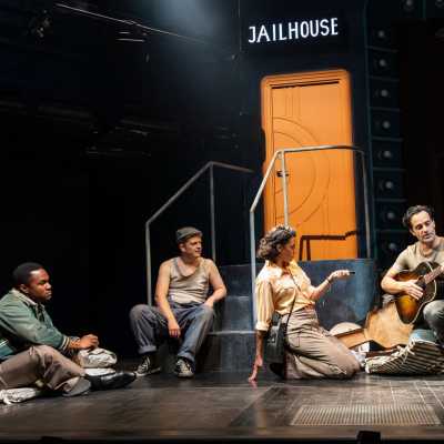 A group of people on a dark stage, under a sign that reads 'Jailhouse'. One of them plays a guitar, while another person holds a small microphone.