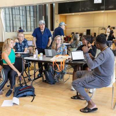 8 members of the cast and creative team are gathered around a table, talking. There is a mirrored wall behind them.