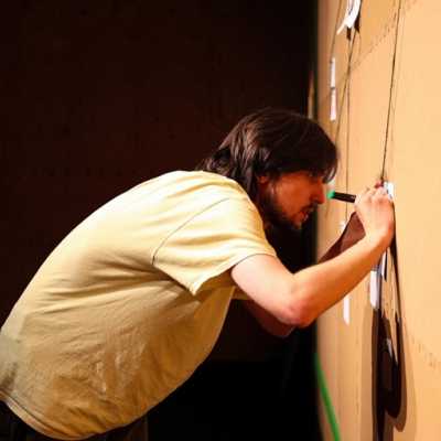 A person in a pale yellow t-shirt viewed from the side writing on a piece of card pinned on a corkboard. 