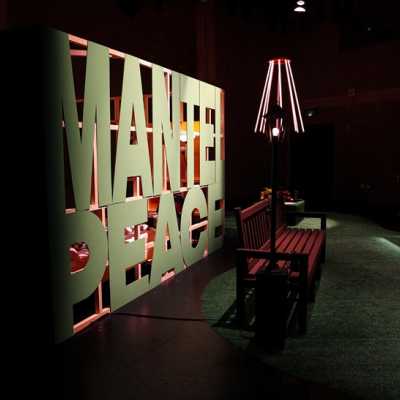 A park scene in a dimly lit studio, with a grassy floor, a park sign, bench and lamppost. Behind the bench, large green letters read "MANTELPEACE". In the background, a light made of multiple warm white LEDs hangs from the ceiling. 