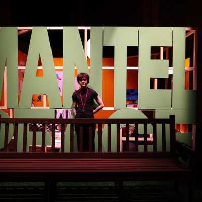 A person dressed in black stands behind a bench in front of large green lettering reading "MANTELPEACE"