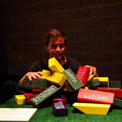 A person smiles at the camera kneeling behind a grassy podium on which sit multi coloured blocks with different positive traits written on them. 