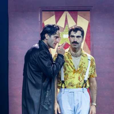 Two people stand in front of a purple doorway. One, who is dark-haired and clean shaven, holds up a finger to an olive-skinned Mediterranean man