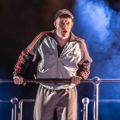 A young man wearing a grey Adidas tracksuit top, grey tracksuit bottoms and a Nike cap is leaning over a metal railing. His mouth is open, mid-speech, and he is wearing a serious expression.  