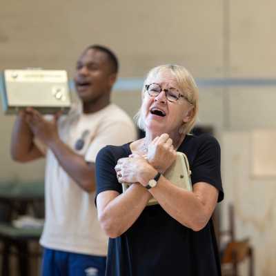 Cast member Annie Wensak is pictured mid-song, clutching a 50s style radio to her chest. In the background, fellow actor Durone Stokes can be seen, also mid-song, holding a radio close to his face.