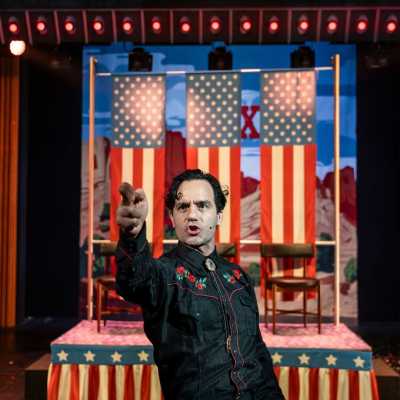 A clean-shaven person with dark hair holds up their fingers like a pistol, in front of a float decorated with the American flag.