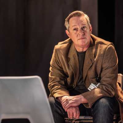 A man in a brown jacket and blue jeans sits on a plastic chair. He is wearing a serious expression and is clasping his hands in front of him, elbows resting on his knees. In front of him is an empty plastic chair. 