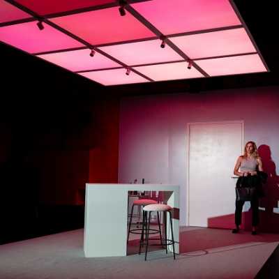 A white Welsh woman with blonde shoulder-length hair stands at the entrance to a modern-looking apartment. The ceiling is made up of pink lights.