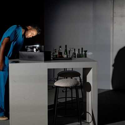 A brown-skinned, non-binary woman with short Afro hair wears blue scrubs and stands at a breakfast bar, peering at an illuminated object on the bar.