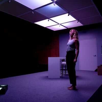 A white, average height Welsh woman with blonde shoulder-length hair dances on a white stage under a ceiling of white panel lights looking at a black box on the floor. She wears a grey vest and black trousers. 