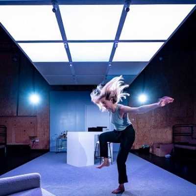 A white Welsh woman with blonde shoulder-length hair dances on a white stage under a ceiling of white and blue lights.