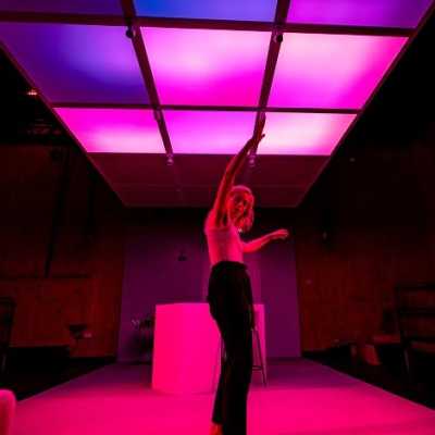 A white Welsh woman with blonde shoulder-length hair stands on a white stage under a ceiling of pink lights with her arms up in the air.