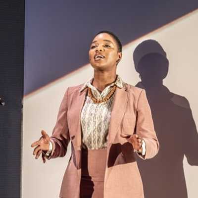 A Black woman of average height with short hair stands singing. She wears a peach coloured suit.