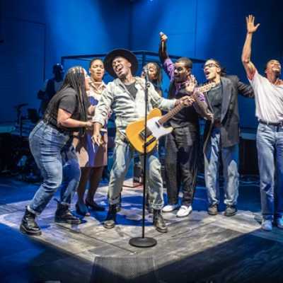 A group of people stand around a microphone, singing. One of them, in the centre, plays an electric guitar.