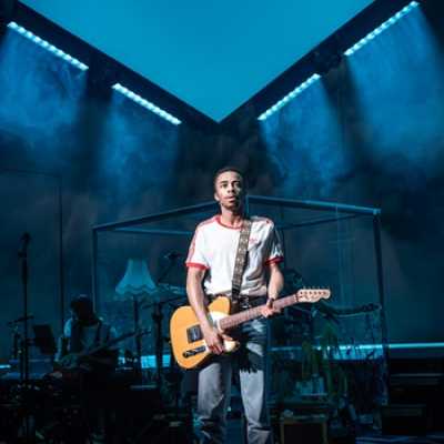 An average height, slim,  Afro-Caribbean man wearing a red and white top and jeans stands holding an electric guitar. Behind him a band is visible, including a glass box containing a drum kit. A white v-shaped ceiling is above him.