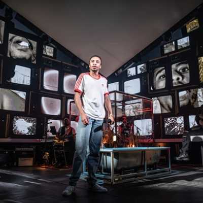 An average height, slim, Afro-Caribbean man wearing a red and white top and jeans stands in front of two large walls of retro tv screens in a V-formation. Each of the tv screens depicts a different image, like a portion of his face. Other figures stand around the area.