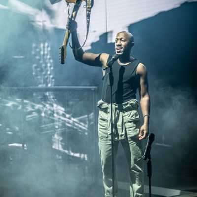 A Black man of average height stands in front of a microphone. He holds an electric guitar above his head with one arm, and wears black vest and boilersuit tied around his waist.