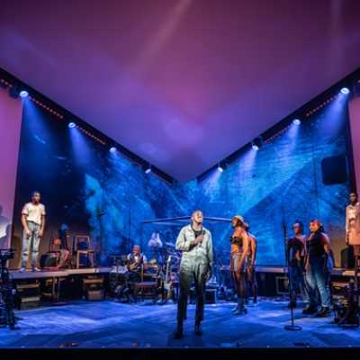 A group of people stand on a wide stage, with light pink walls and ceiling, and walls portraying blurred video images. There are amps and instruments littered around the stage, including a drum kit and guitars. There are also chairs and other props dotted around the stage.