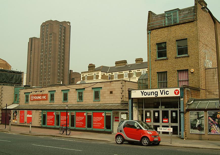 A photo of the Young Vic before it's rebuild in 2006. The old red YV branding covers the walls of the low slung building with the old house that was once a bakery at the East end of the building. A red Smart car sits outside the box office entrance. 