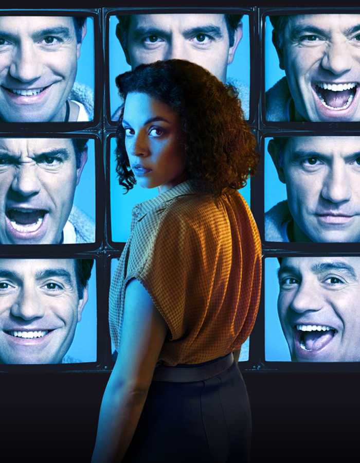 A series of 9 TV screens stacked in columns of 3 show a person's face with different expressions on a blue background. In front of the TV screens stands a person with shoulder length curly hair wearing a yellow blouse turning over their left shoulder to look towards the camera. 