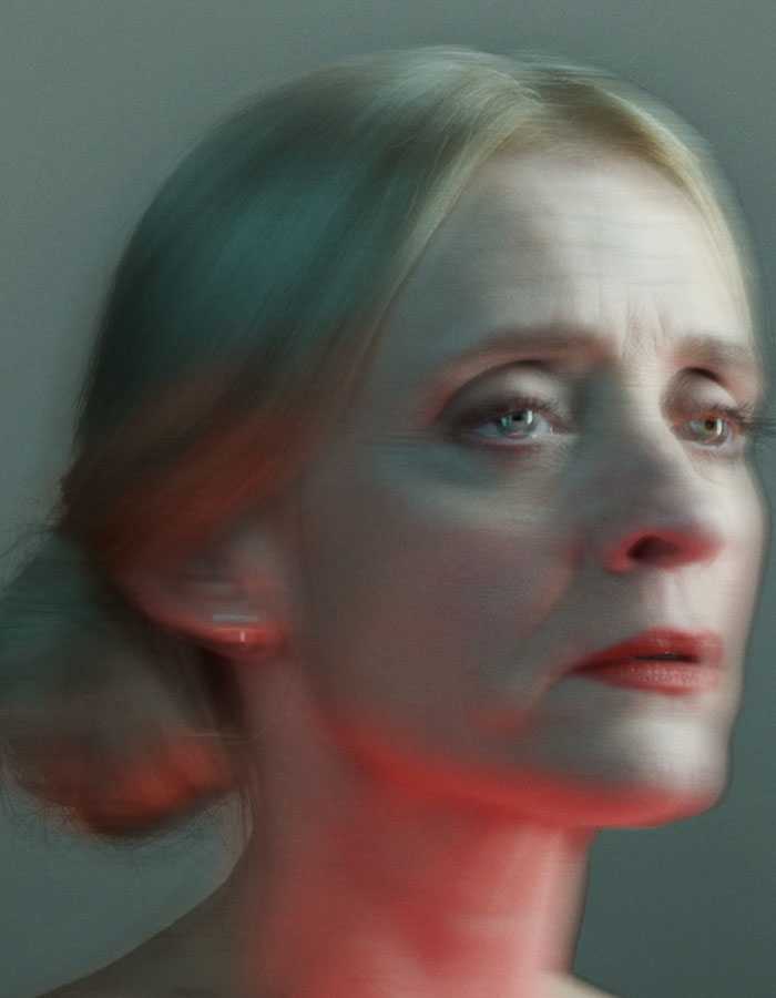 A blurred image of the head and shoulders of Anne-Marie Duff. Her blonde hair is pulled into a bun and her neck is lit from below with a red light. 