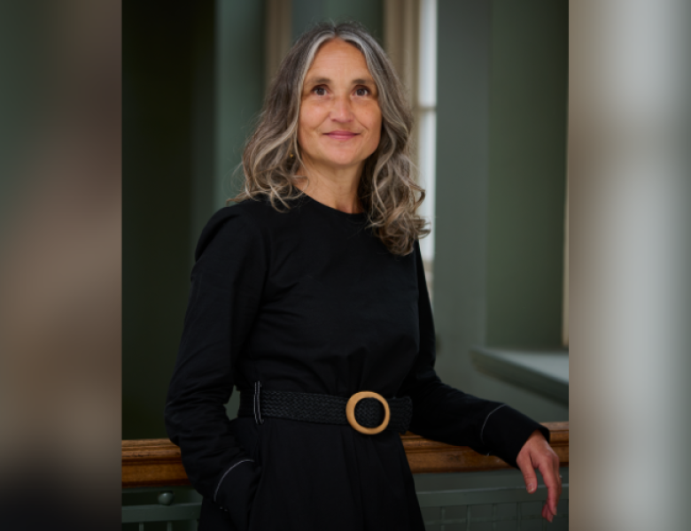 A white woman with shoulder length wavy brown and grey hair, and dark eyes, wearing a black dress.